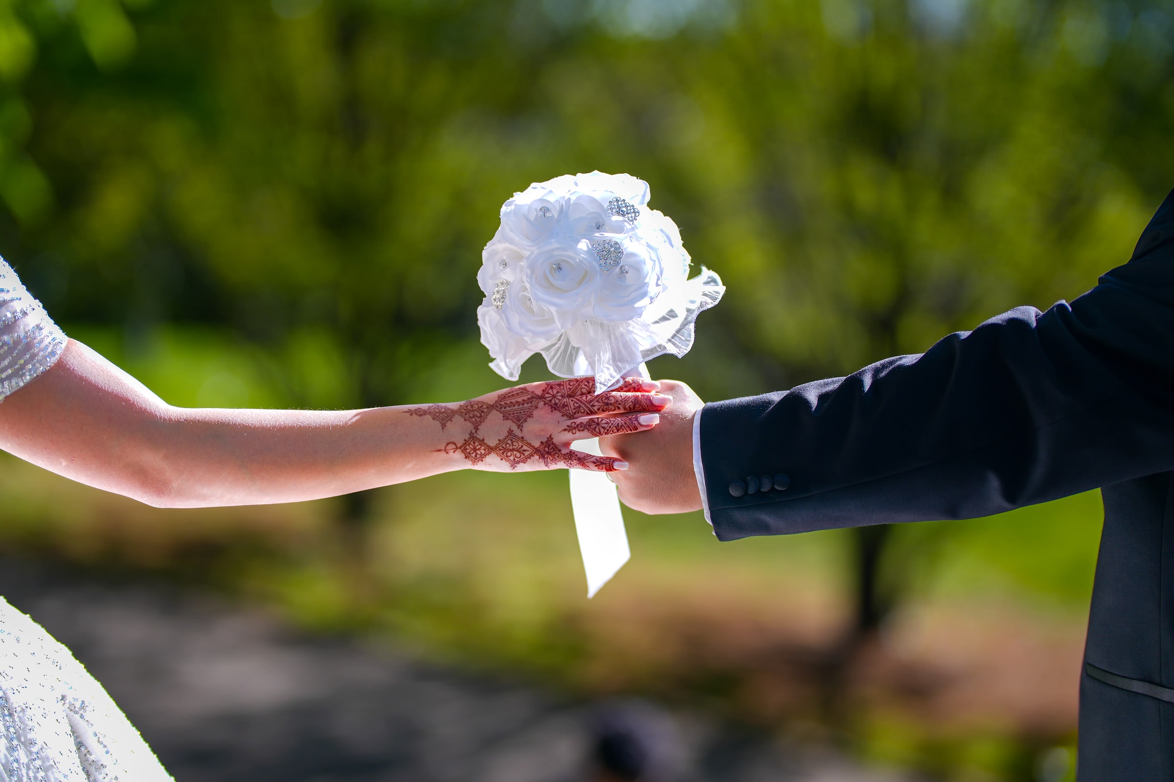 Les mains des mariés se rejoignent autour d’un bouquet blanc, dans un parc